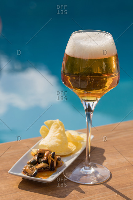 Fresh cold beer in goblet served with mussels and lemon on wooden counter against swimming pool in summer day
