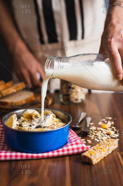 Crop anonymous housewife pouring yogurt from glass bottle into bowl with cereals and banana while preparing healthy breakfast at home kitchen