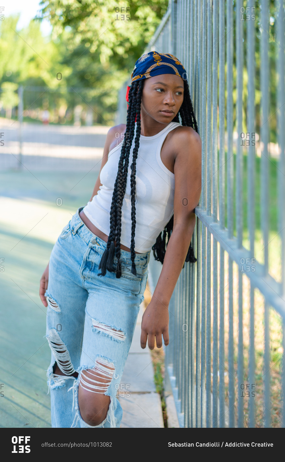 Young African American female with Afro pigtails dressed in stylish ripped jeans and white top leaning on metal fence in urban park and looking away