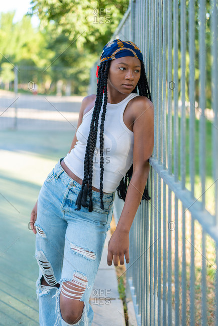 Young African American female with Afro pigtails dressed in stylish ripped jeans and white top leaning on metal fence in urban park and looking away