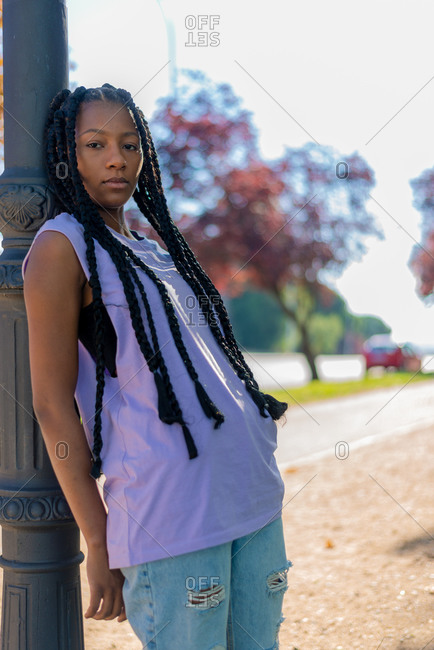 Serious youthful African American female with braids wearing casual shirt and ripped jeans leaning on pillar and looking at camera in urban park in summer day
