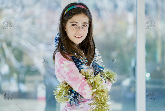 Positive school aged girl in casual wear wrapped in colorful shiny tinsel looking at camera while standing against window at home during Christmas preparation