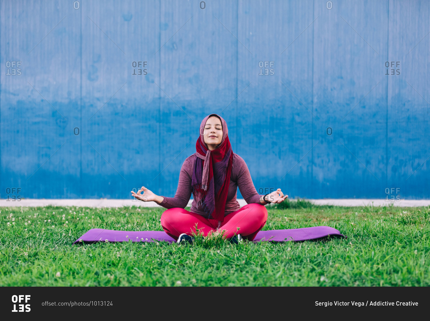 Serene Arab female in sportswear and hijab sitting on mat on lotus pose with eyes closed in park and doing yoga in Padmasana with closed eyes