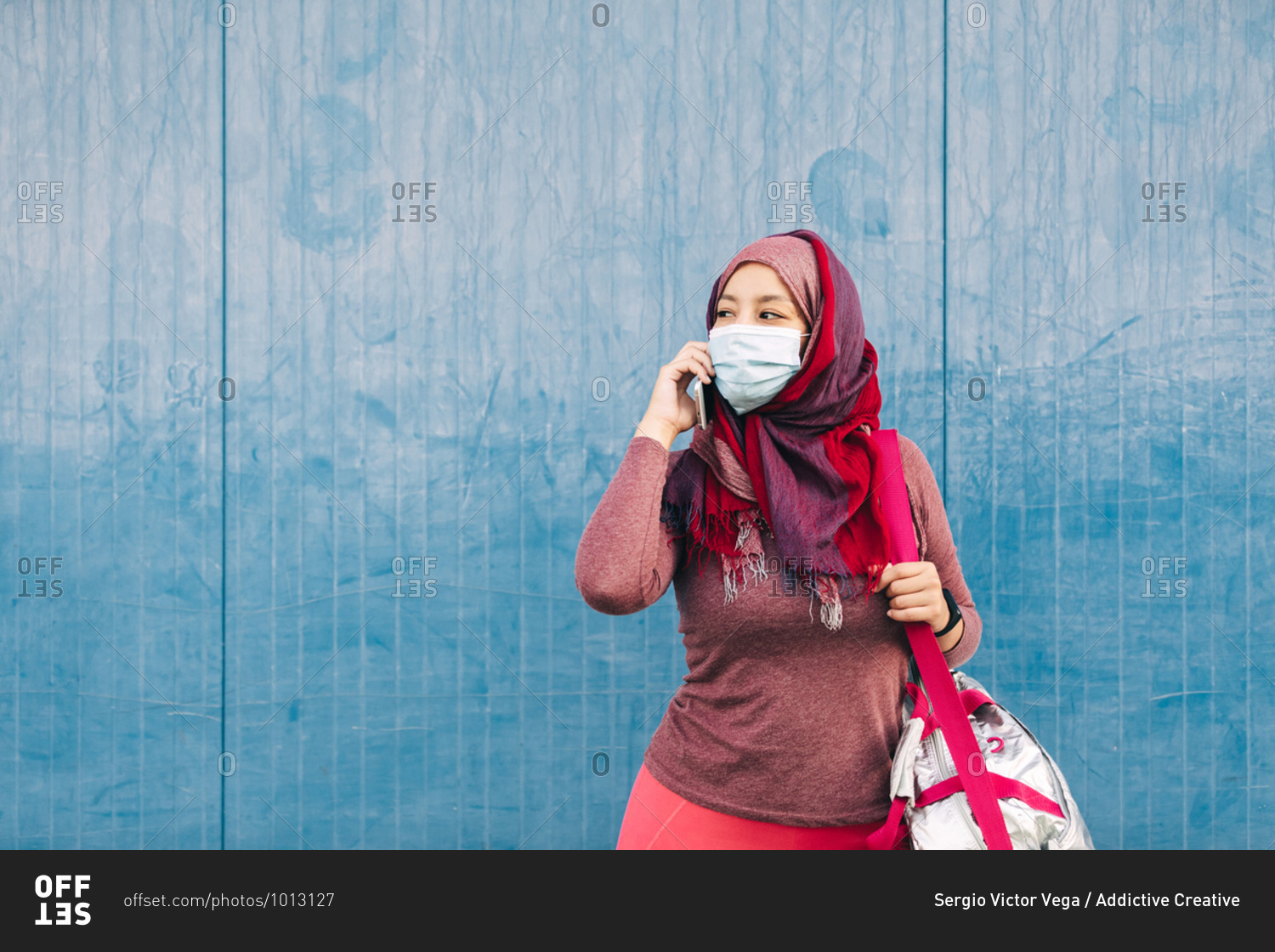 Arab female in sportswear and medical mask standing near building in city speaking on mobile phone after training while looking away