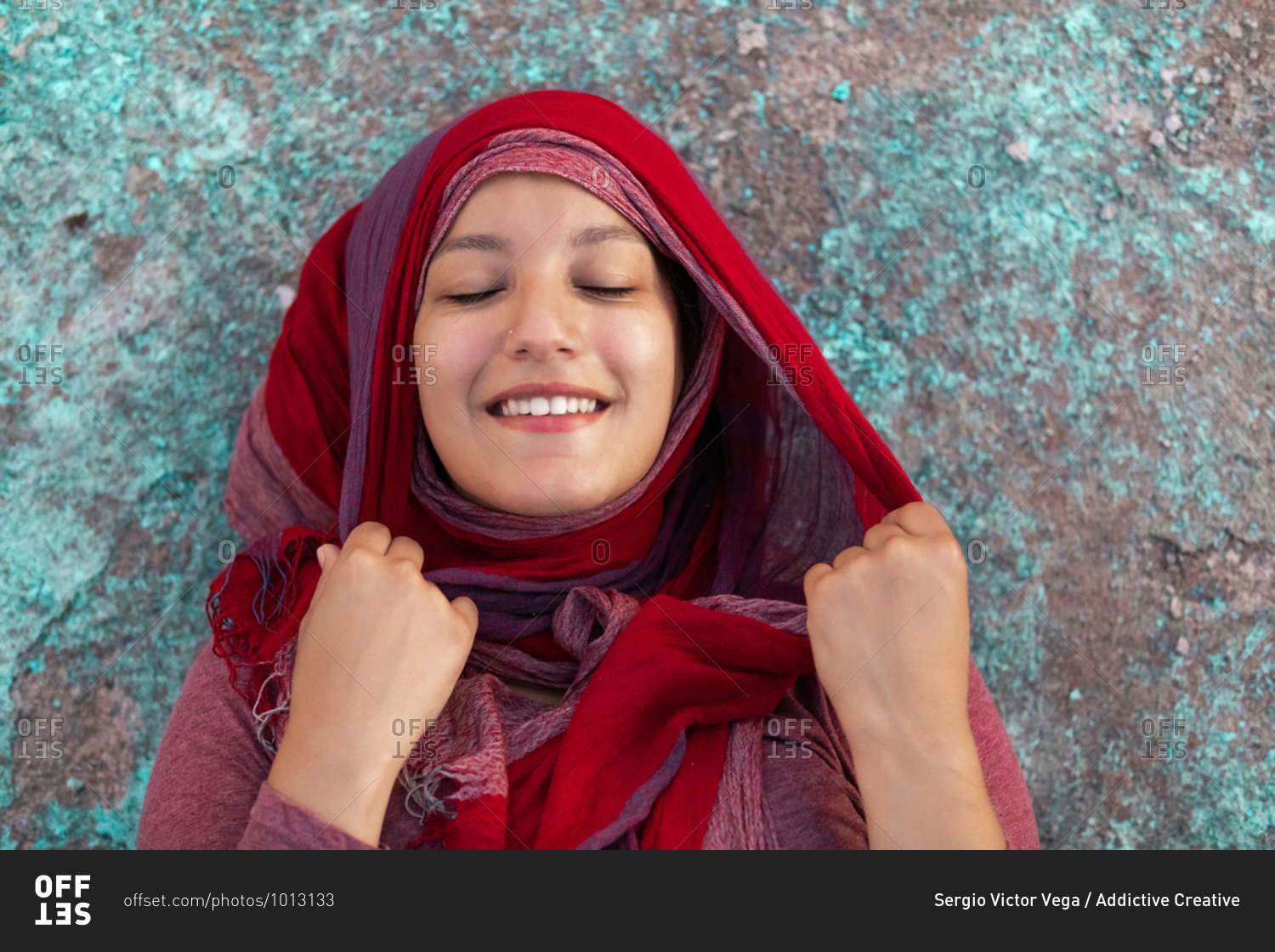 Happy Arab female wearing hijab leaning on concrete colorful building in city smiling with eyes closed