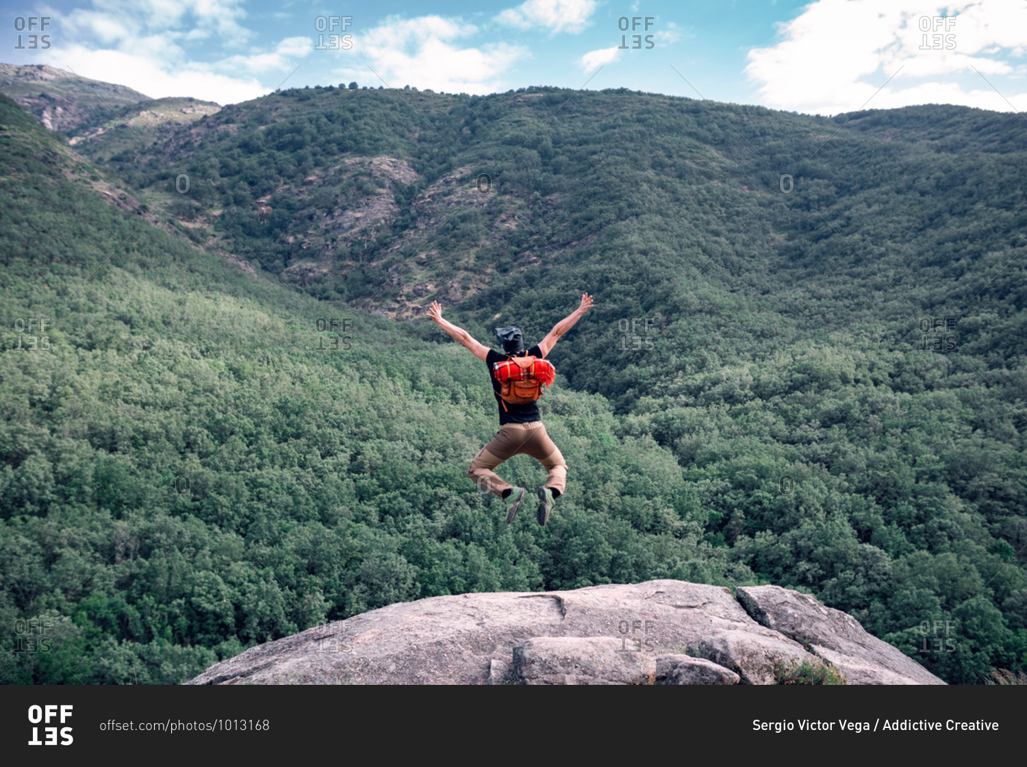 Back view of faceless male hiker with trekking poles and backpack in moment of jumping with outstretched arms on background of mountainous landscape