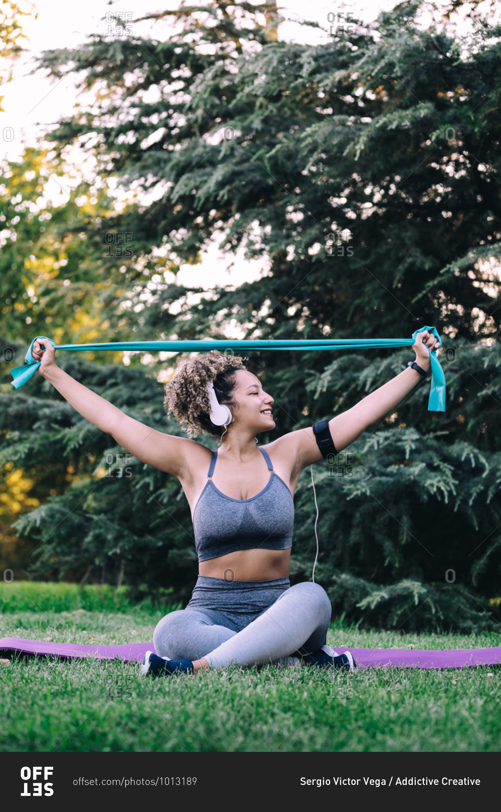 Full body positive young fit female with headphones sitting with legs crossed on grass and stretching arms with resistance band during fitness training in summer park