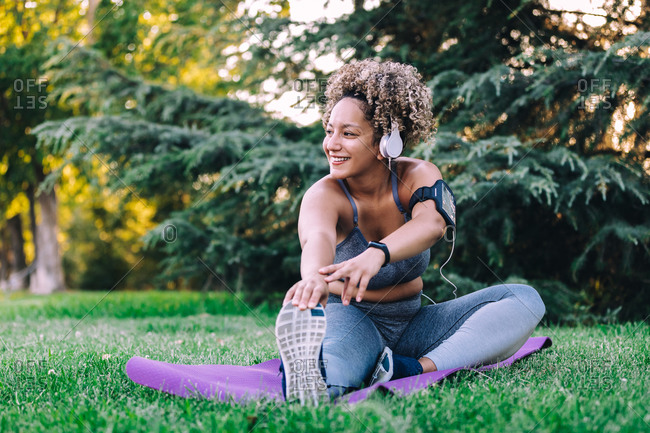 Happy young ethnic female with curly hair in sportswear listening to music with headphones and smartphone and doing seated forward bend exercise while stretching body during fitness training in summer park