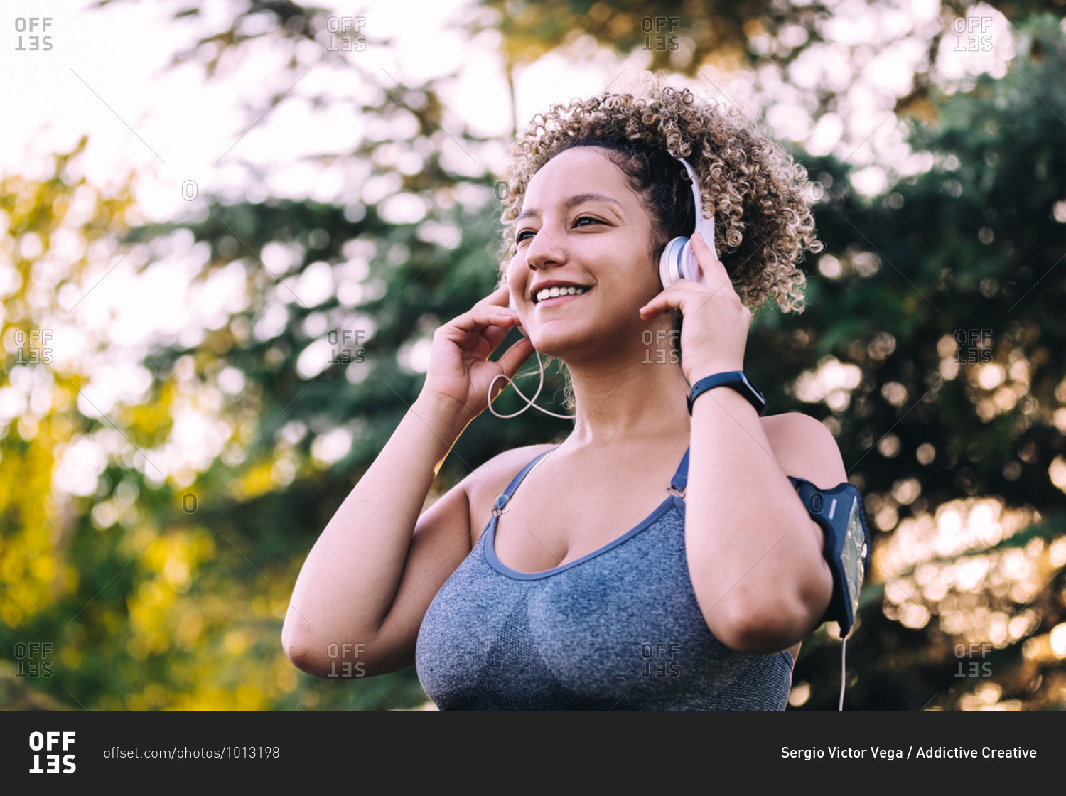Low angle of optimistic young ethnic female with Afro hairstyle adjusting headphones while listening to music on smartphone during training in green park