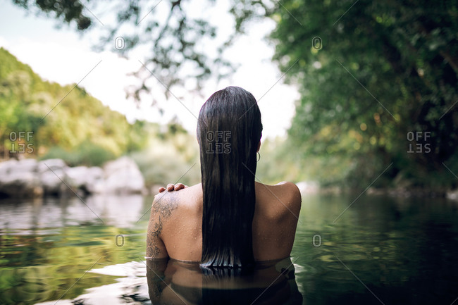 Back view of unrecognizable female with bare shoulders and wet hair standing in water of calm pond in woods and enjoying summer vacation
