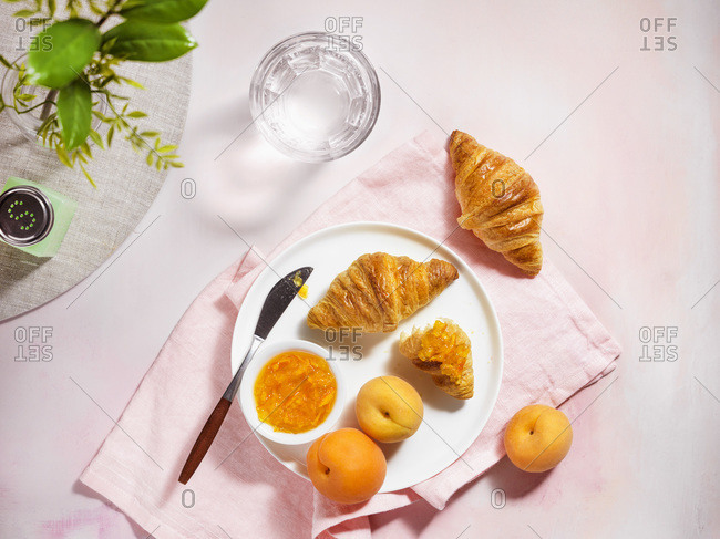 A top down view of a bright, summer breakfast scene with croissants, apricots and jam on a pale pink, textured tabletop.