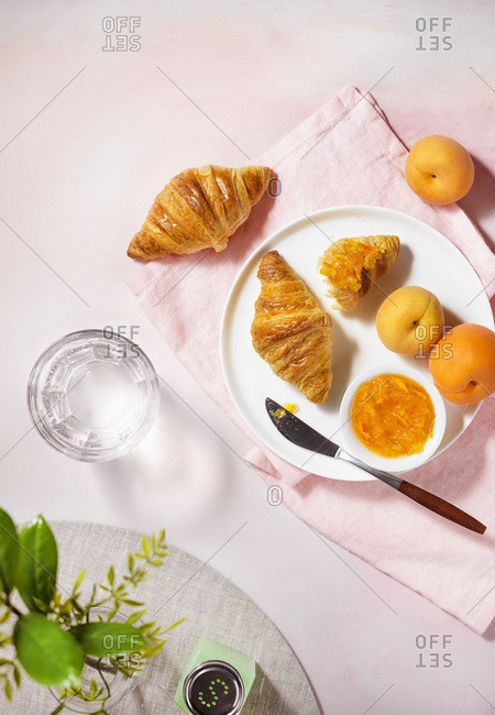 A top down view of a bright, summer breakfast scene with croissants, apricots and jam on a pale pink, textured tabletop.