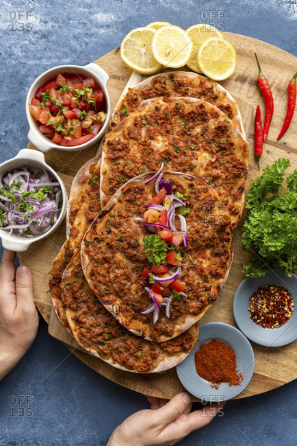 Woman holding a wooden board to serve homemade Turkish lahmacun with red onion salad and tomato salad as condiments. Spices, lemon slices, a bunch of parsley and chili pepper accompany.