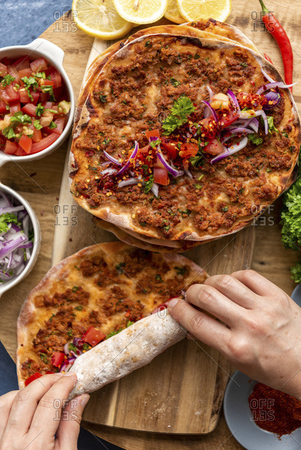 Hands rolling up a lahmacun on a wooden board with some more lahmacun portions and condiments like red onion salad, tomato salad, lemon slices, chili pepper and parsley.
