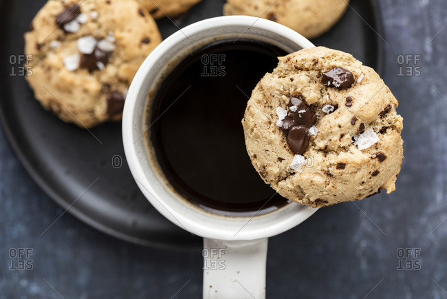 A tahini cookie with chocolate chunks put on the edge of a white ceramic mug filled with dark coffee on a black plate.