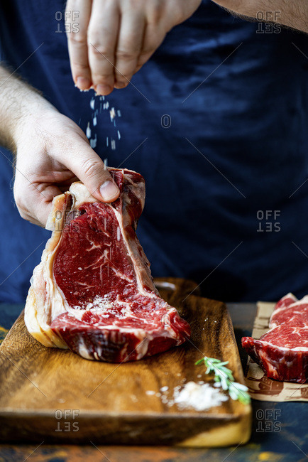 Man sprinkling salt over a raw rib eye steak on a wooden board. A pinch of salt and a sprig of rosemary on the board accompany.