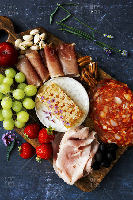 Meat platter with honey comb, chorizo, roast beef, prosciutto, grapes, pistachios, strawberries, pecans and olives on a dark background.