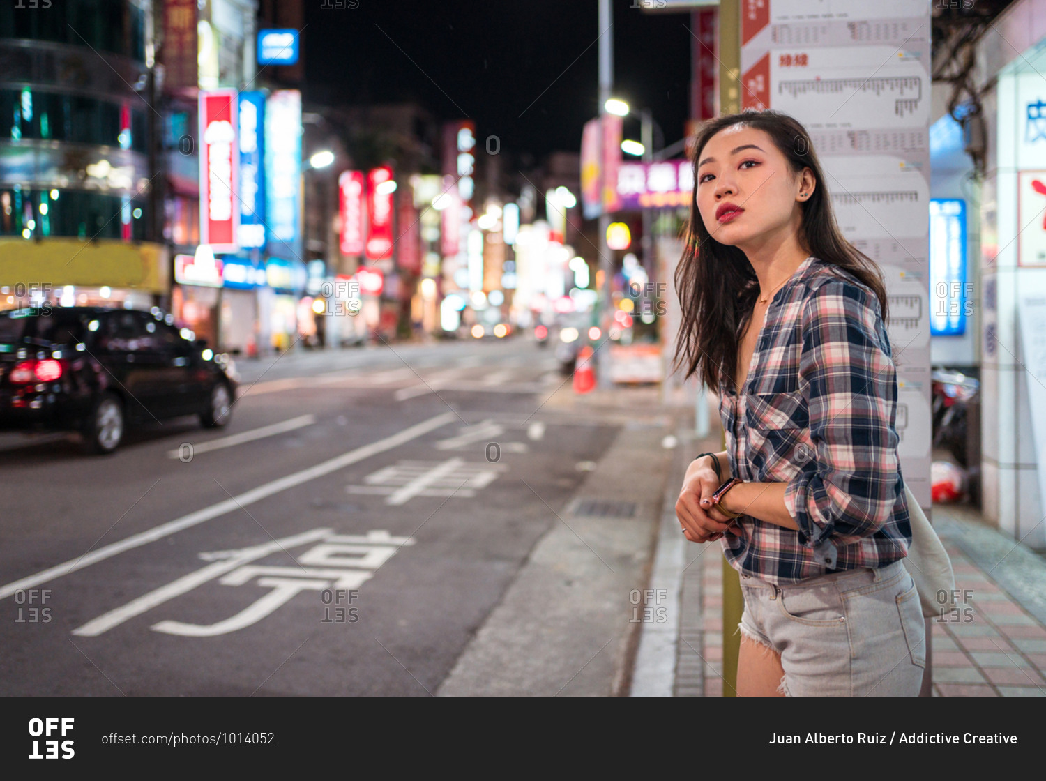 Side view of young Asian female in casual outfit looking away with hope while waiting for urban bus on bus stop in city during night time