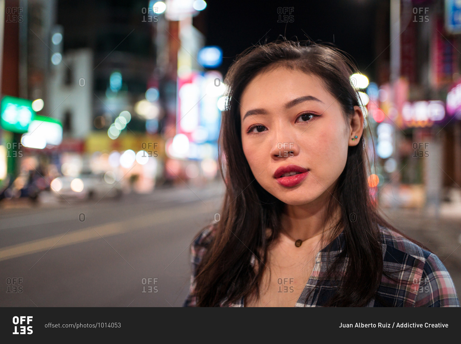 Millennial Asian female in checkered shirt looking at camera with smile while crossing street in modern illuminated city district at night time