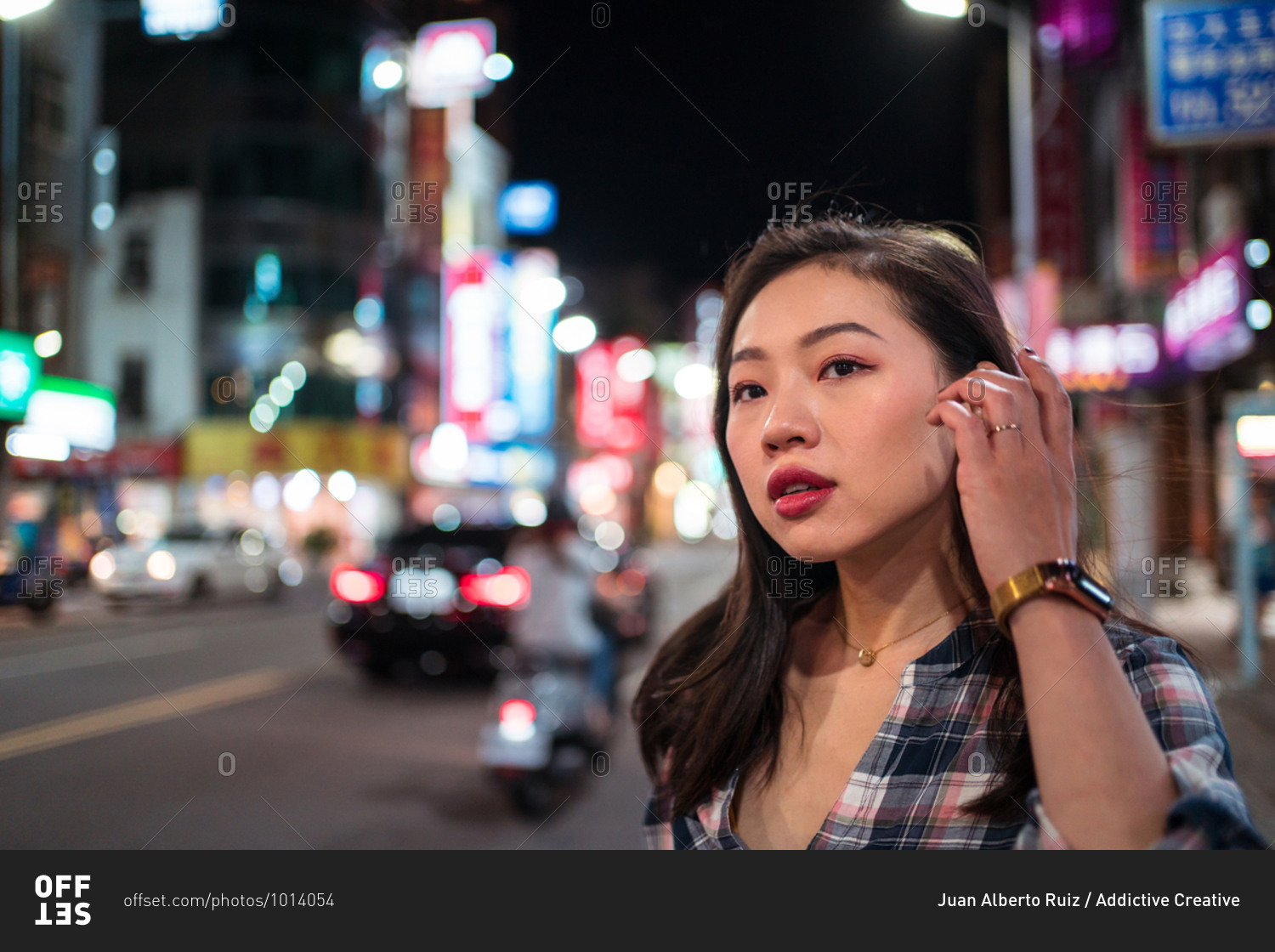 Millennial Asian female in checkered shirt looking away with smile while crossing street in modern illuminated city district at night time