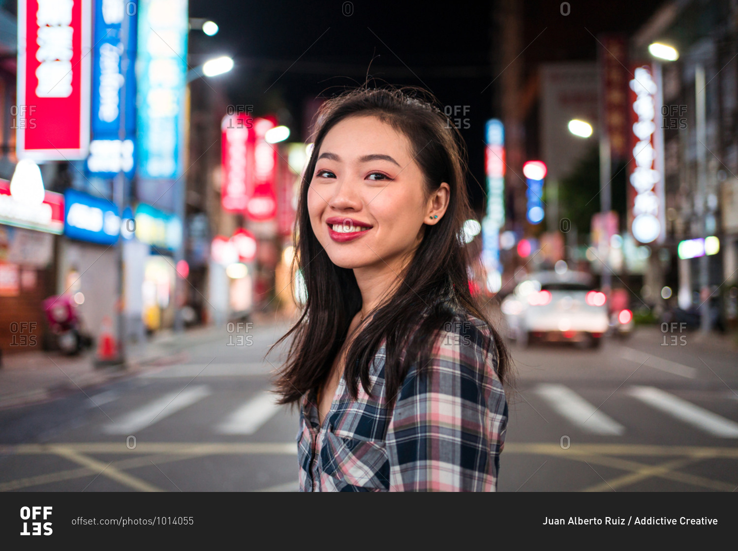 Side view of cheerful millennial Asian female in checkered shirt looking at camera with smile while crossing street in modern illuminated city district at night time
