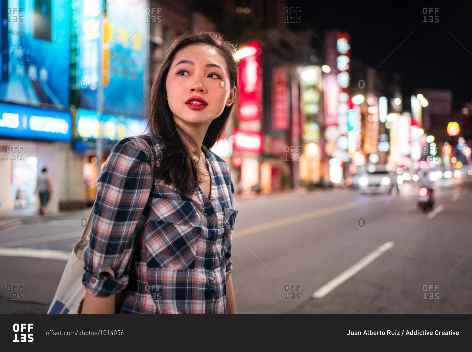 Side view of cheerful millennial Asian female in checkered shirt looking away while crossing street in modern illuminated city district at night time