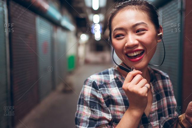 Joyful young Asian female student in casual clothes communicating with friends through headset while standing on blurred urban background
