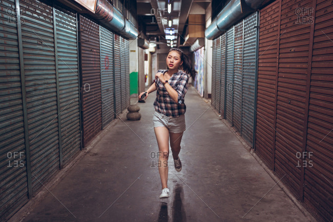 Full body young Asian female in casual clothes with smartphone in hand running fast in empty underground corridor with closed shutters