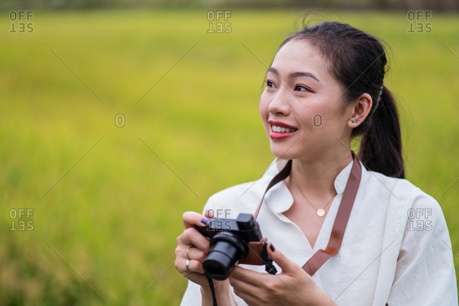Delighted Asian female in summer wear standing in rice field and taking pictures on vintage photo camera