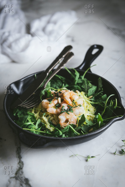 From above portion of delicious zucchini noodles with boiled prawns and fresh spinach leaves placed on frying pan with fork and knife on marble table