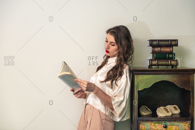 Side view of positive young female in stylish outfit leaning back on vintage cabinet and reading interesting book in cozy room