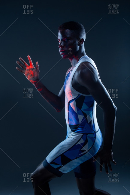 Side view of serious athletic African American sportsman in colorful active wear performing running fast movement in dark studio with red neon illumination