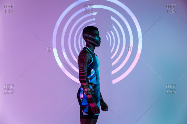 Side view of determined muscular young African American sportsman standing against wall with concentric circles in studio with neon lights