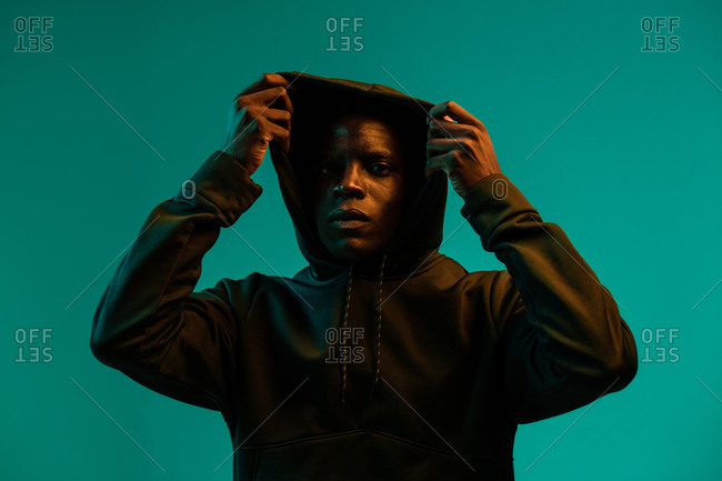 Serious young African American male in black hoodie covering head with hood against green background in studio