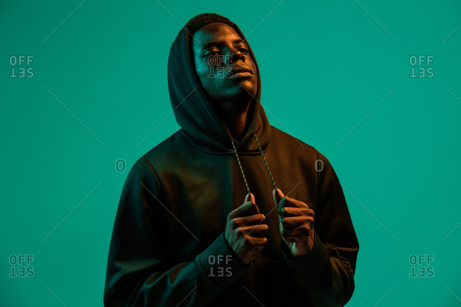 Serious young African American male in black hoodie covering head with hood against green background in studio looking away