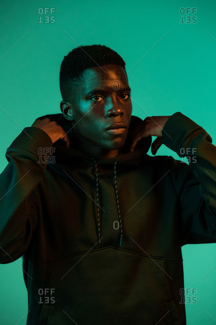 Serious young African American male wearing a black hoodie covering head with hood against green background in studio