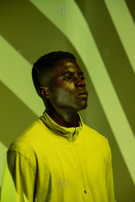 Young African American male athlete in sportswear looking away while standing under green neon illumination near corner with striped shadows in studio
