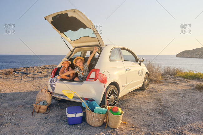 Two children sitting inside the trunk of the car with gestures of freedom with baskets of objects for the beach and a portable cooler next to the car with the sea in the background