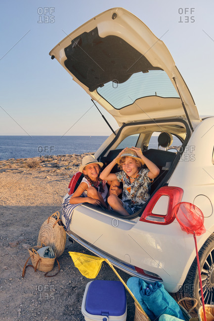 Vertical photo of two children sitting inside the trunk of the car making joke gestures with baskets of objects for the beach and a portable cooler next to the car with the sea in the background