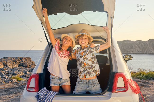 Two children bending inside the trunk of the car with their hands raised in a gesture of victory while wearing a straw hat with baskets of objects for the beach next to the car with the sea in the background