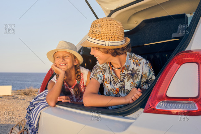 Blonde girl inside the trunk of a white car making a wait gesture with a happy expression next to a boy in a straw hat and the sea in the background