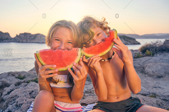 Two children playing with a piece of watermelon imitating a smile with it sitting on some rocks in front of the sea during the sunset