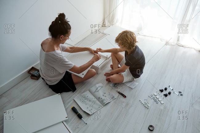 From above of young female with son using paper instruction and fixing details of wooden wardrobe while assembling furniture together in new home