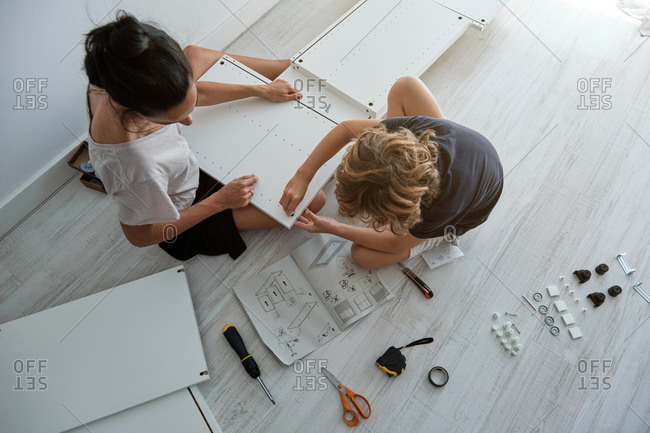 From above of young female with son using paper instruction and fixing details of wooden wardrobe while assembling furniture together in new home