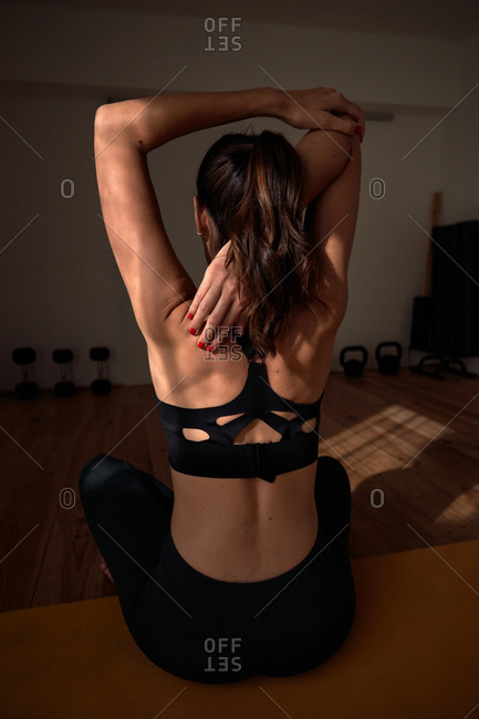 Back view of anonymous female sitting with crossed legs on mat and stretching arms behind back while practicing yoga
