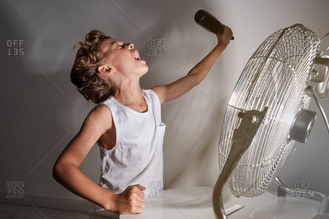 Child with a white shirt with a raised arm holding a hairbrush that mimics a microphone singing in front of a running fan in a room