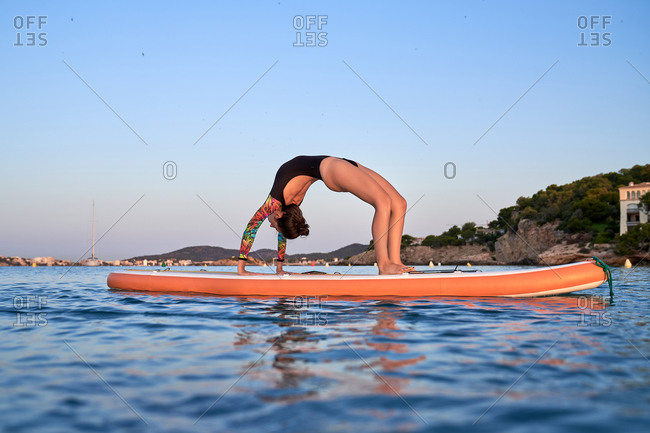 Side view of unrecognizable female in swimsuit standing in Urdhva Dhanurasana on paddleboard in sea and doing yoga during sundown