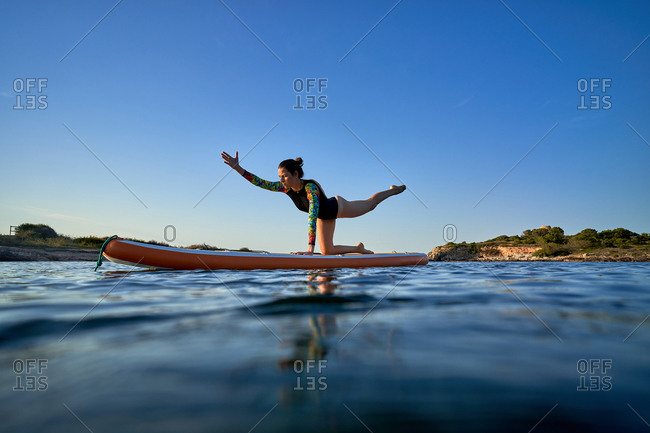 Concentrated female surfer practicing yoga in Utthita Cakravakasana while balancing on paddleboard in sea during sunset