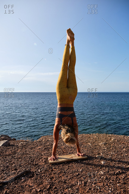 Vertical photo of a woman in yellow tights practicing handstand yoga with her feet up and her hands resting on a wooden board in front of the sea
