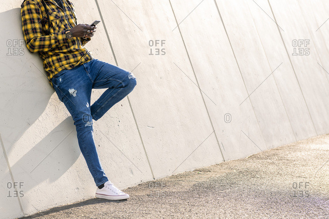 Unrecognizable young black man writing a message on his phone leaning on a modern concrete structure, concept of lifestyle and technology